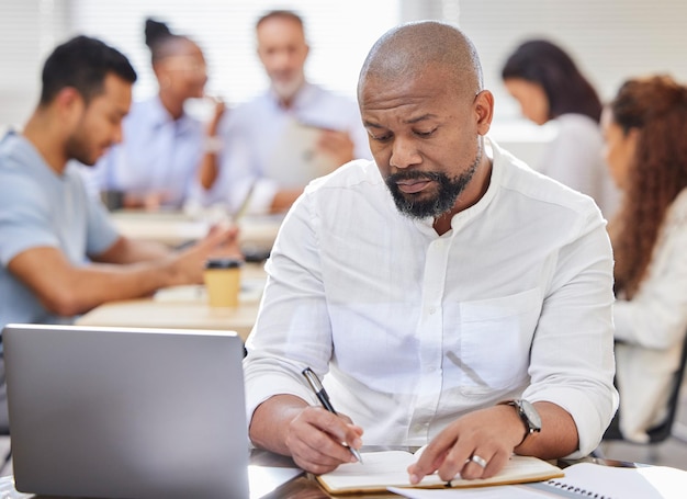 Photo l'homme en charge des grands projets photo d'un homme d'affaires mature écrivant des notes tout en travaillant sur un ordinateur portable dans un bureau avec ses collègues en arrière-plan