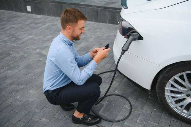 Homme chargeant sa voiture électrique à la station de charge et utilisant un smartphone