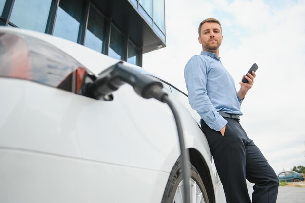 Homme chargeant sa voiture électrique à la station de charge et utilisant un smartphone