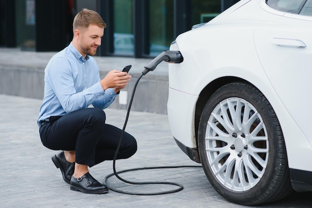 Homme chargeant sa voiture électrique à la station de charge et utilisant un smartphone