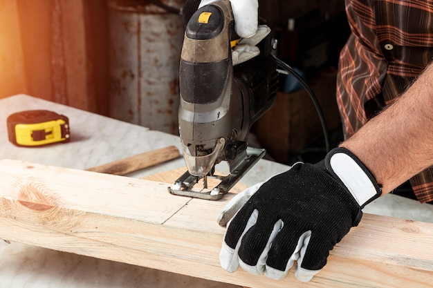 Un homme charpentier coupe une poutre en bois en utilisant un puzzle électrique, mains mâles avec un gros plan de puzzle électrique.