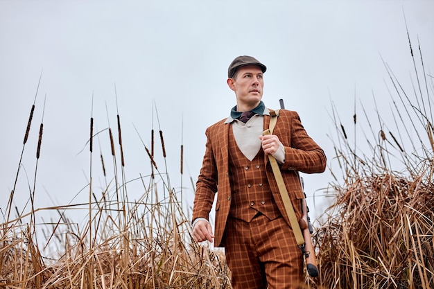 Homme de chasseur caucasien américain confiant avec fusil de chasse marchant dans la nature de la campagne, portant un costume et un chapeau élégants à la mode marron. chasseur professionnel qualifié et expérimenté en attente de proie