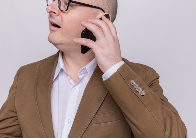 Homme chauve d'âge moyen en costume portant des lunettes souriant heureux et positif tout en parlant au téléphone mobile