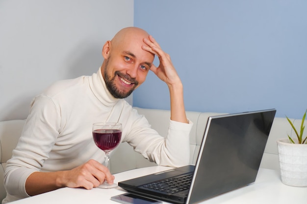 Homme chauve avec barbe en col roulé blanc, boire du vin utiliser un ordinateur portable à la maison en souriant à la caméra