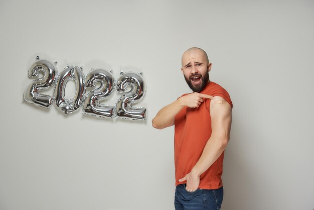 Un homme chauve avec une barbe crie et montre un pansement médical sur son épaule après la vaccination contre le coronavirus (Covid-19) près de ballons d'argent déjoués en forme de 2022.