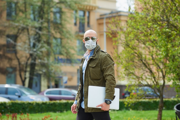 Un homme chauve avec une barbe dans un masque médical pour éviter la propagation du coronavirus se promène avec un ordinateur portable dans le parc. Un gars porte un masque facial n95 et des lunettes de soleil pilotes dans la rue de la ville.