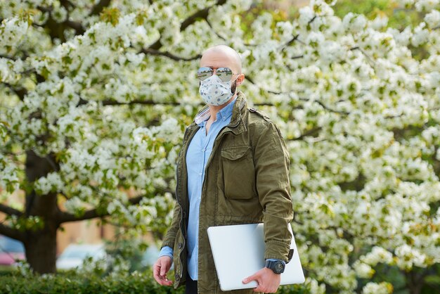 Un homme chauve avec une barbe dans un masque médical pour éviter la propagation du coronavirus se promène avec un ordinateur portable dans le parc. Un gars porte un masque n95 et des lunettes de soleil pilotes dans le jardin entre les arbres en fleurs.