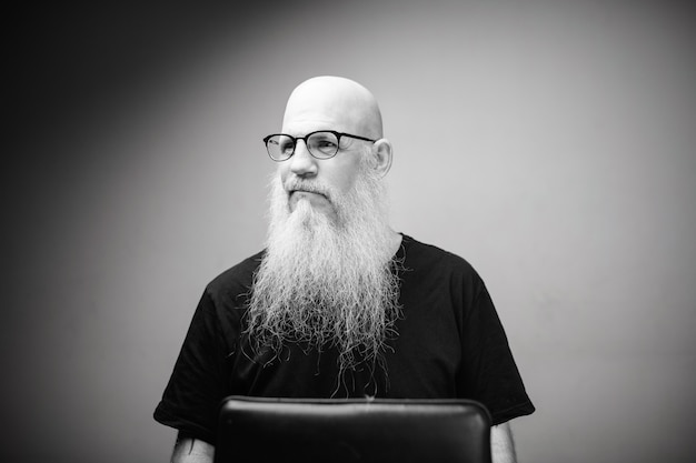 Homme chauve mature avec une longue barbe grise contre un mur blanc en noir et blanc