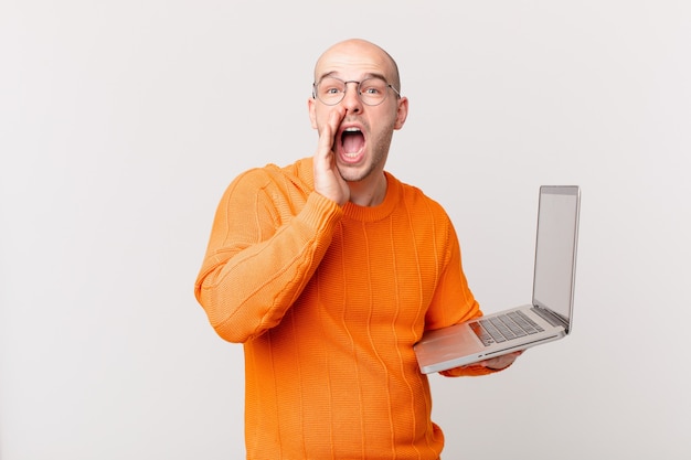 Homme chauve avec ordinateur se sentant heureux, excité et positif, donnant un grand cri avec les mains à côté de la bouche, appelant