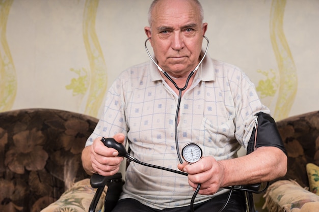 Homme chauve senior ayant des besoins spéciaux, assis sur son fauteuil roulant et tenant un sphygmomanomètre et un stéthoscope, regardant directement la caméra.