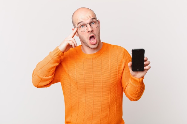 Homme chauve avec un smartphone ayant l'air surpris, bouche bée, choqué, réalisant une nouvelle pensée, idée ou concept