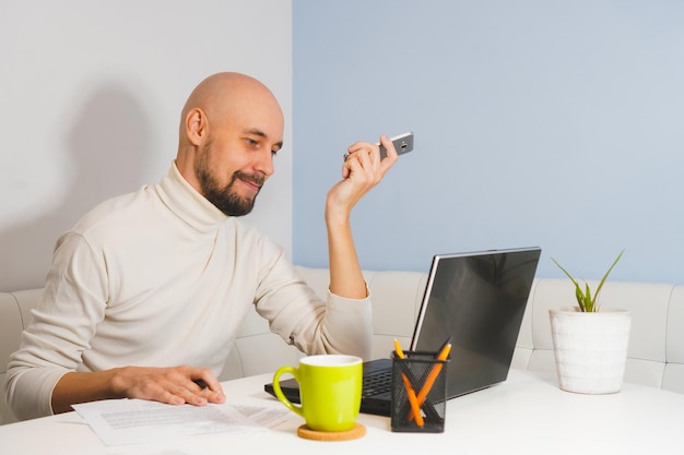 Homme chauve souriant avec barbe en col roulé blanc travaillant à la maison sur un ordinateur portable tenant un téléphone