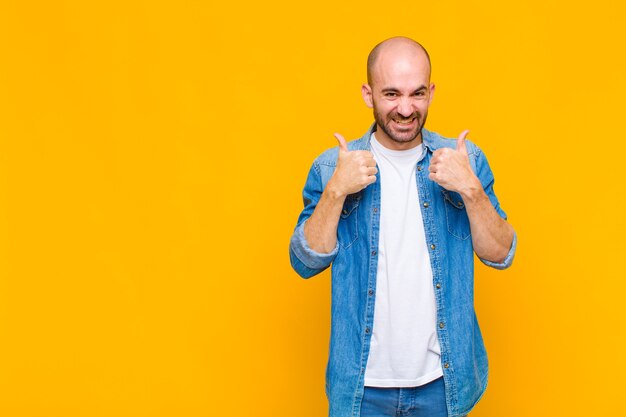 Homme chauve souriant largement à la recherche de bonheur, positif, confiant et réussi, avec les deux pouces vers le haut