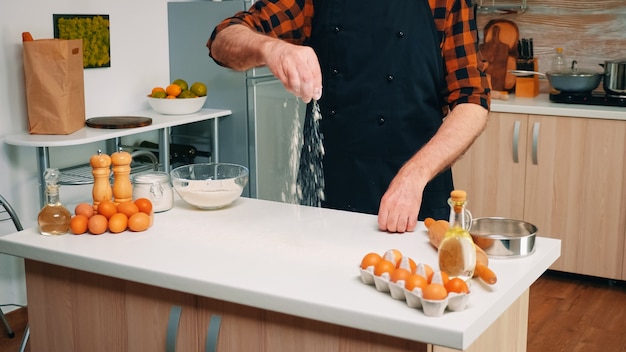 Homme chef principal prenant la farine d'un bol en verre et tamisant sur la table. Boulanger senior à la retraite avec bonete et saupoudrage uniforme, tamisant, étalant des ingrédients rew cuisant des pizzas et du pain faits maison.