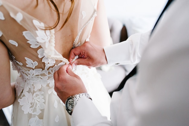 Un homme en chemise blanche avec une cravate et une montre ferme des boutons sur le corset de la robe. Mariée en robe de mariée avec de la dentelle debout dans la chambre.