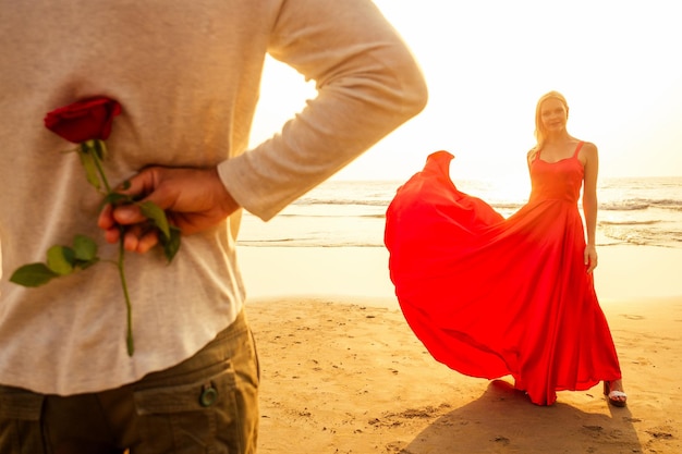 Homme en chemise blanche donnant des roses à une merveille surprise dans une longue robe rouge sur la plage de l'océan mariage romantique ou concept de la Saint-Valentin par un couple amoureux célébrant la journée de la femme du 8 mars