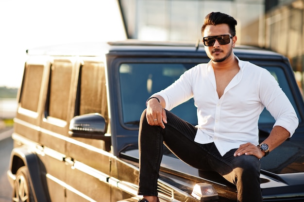 Homme en chemise blanche et lunettes de soleil posant près de voiture suv noir