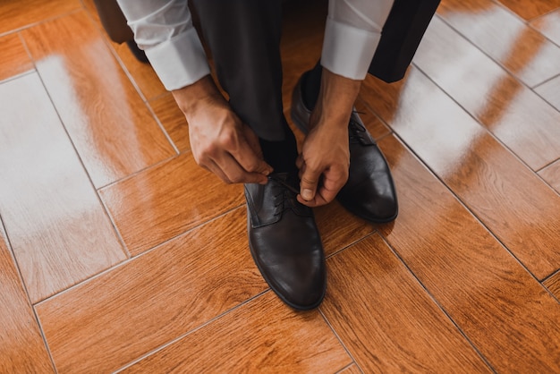 Un homme en chemise blanche en pantalon foncé classique resserre les lacets de ses chaussures sur un sol carrelé noir.