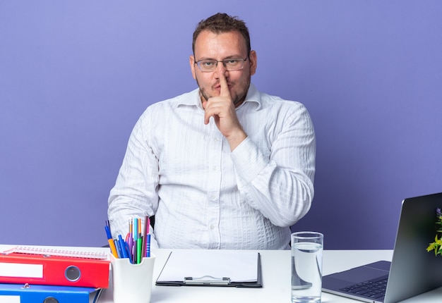 Homme En Chemise Blanche Portant Des Lunettes Faisant Un Geste De Silence Avec Le Doigt Sur Les Lèvres Assis à La Table Avec Un Ordinateur Portable Et Des Dossiers De Bureau Sur Un Mur Bleu Travaillant Au Bureau