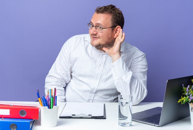 Homme en chemise blanche portant des lunettes regardant de côté heureux et positif avec la main sur l'oreille essayant d'écouter assis à la table avec un ordinateur portable et des dossiers de bureau sur un mur bleu travaillant au bureau