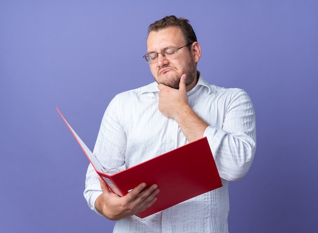 Homme en chemise blanche portant des lunettes tenant un dossier de bureau le regardant avec une expression pensive avec la main sur son menton debout sur le mur bleu