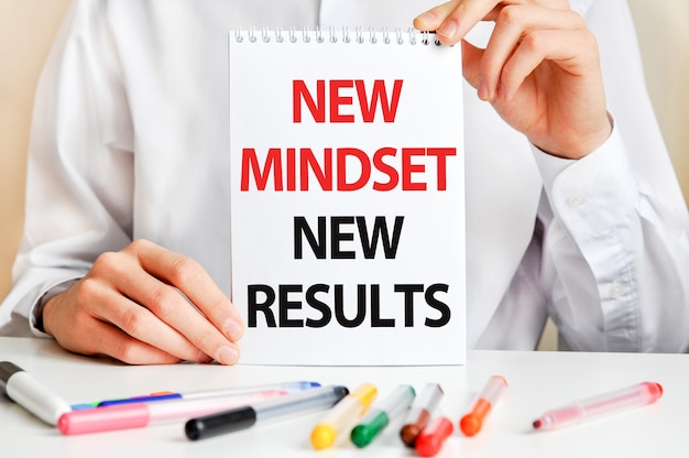 Un homme en chemise blanche tient une feuille de papier avec le texte: NEW MINDSET NEW RESULTS. Marqueurs multicolores et tablette sur une table. Concept commercial et éducatif.