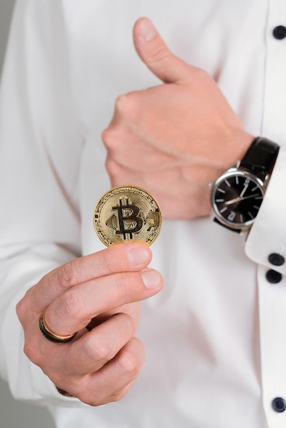 Un homme en chemise blanche tient une pièce d'or bitcoin dans sa main et montre un geste du pouce levé