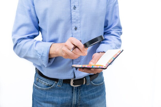 Homme en chemise bleue et pantalon bleu examinant un cahier en papier avec une loupe. Photo sur fond blanc. Espace de copie.