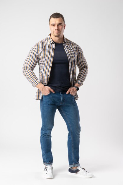 Un homme en chemise à carreaux, chemise bleue et jeans se tient avec les mains sur les hanches.