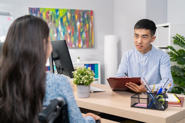 Un homme en chemise est assis derrière un bureau avec un bloc-notes à la main, le patron demande à une femme d'utiliser un fauteuil roulant