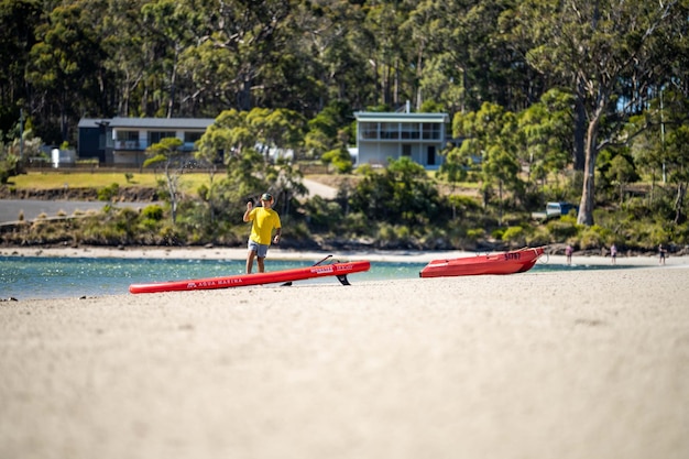 Un homme en chemise jaune se tient sur une plage à côté d'un kayak rouge.