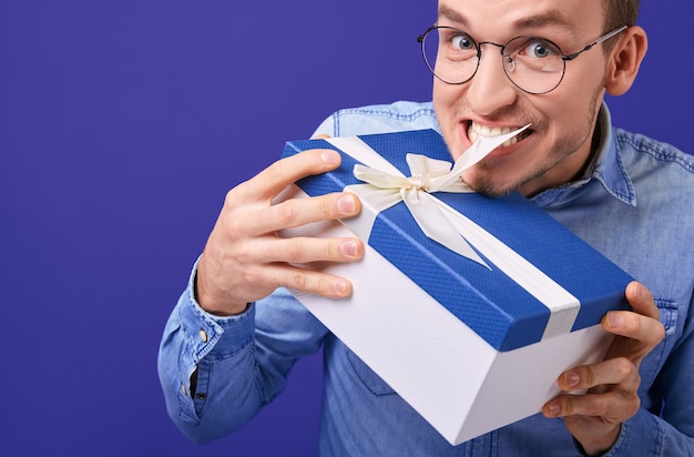 Homme en chemise en jean bleue et lunettes essayant de détacher le cadeau avec ses dents