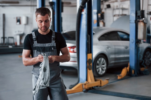 Homme en chemise noire et uniforme gris se tient dans le garage après avoir réparé une voiture cassée