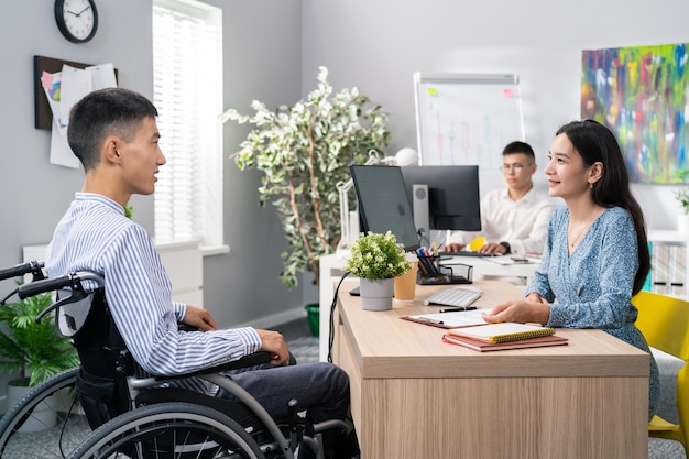 Homme en chemise qui utilise un fauteuil roulant assis à un bureau en face de lui belle femme en robe pose une question