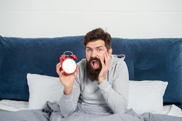 Photo homme choqué avec la mâchoire tombée tenant au réveil dans son lit le matin en train de dormir trop longtemps