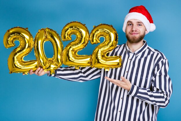 Un homme coiffé d'un chapeau de Noël tient les chiffres 2022 à partir de ballons gonflables