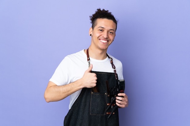 Homme de coiffeur dans un tablier sur fond violet isolé donnant un geste de pouce en l'air