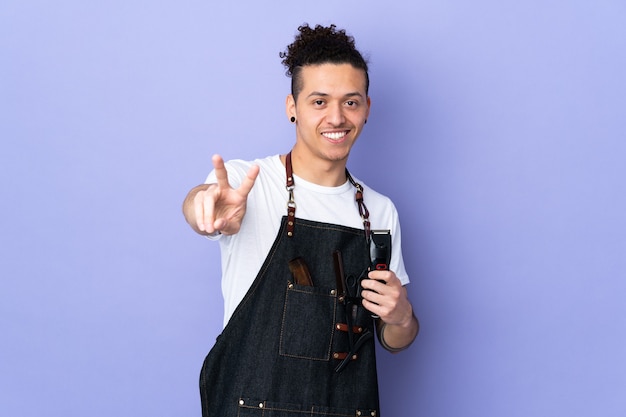 Homme de coiffeur dans un tablier sur fond violet isolé souriant et montrant le signe de la victoire