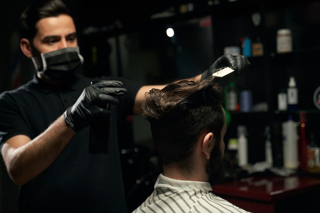 Homme coiffeur professionnel vêtu d'une chemise noire, portant un masque sur la bouche et des gants sur les mains fait la coiffure à un homme non rasé recouvert d'une cape rayée assis dans un fauteuil au salon de coiffure