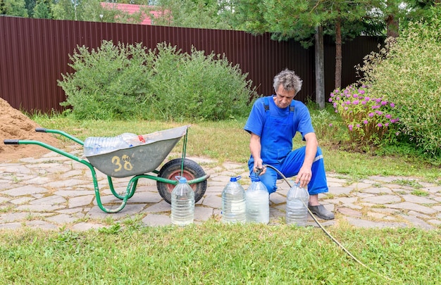 Un homme en combinaison bleue verse de l'eau potable d'un tuyau dans de grandes bouteilles. Eau potable écologique d'un puits dans une maison de campagne.