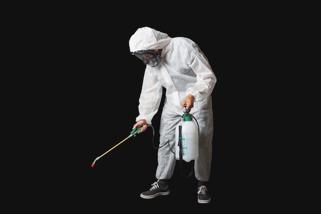 Homme en combinaison de protection blanche avec une capuche portant un masque de baskets et des lunettes de protection tenant un réservoir de pulvérisation avec une buse prête à désinfecter ou à fumiger Le fond est noir massif mettant en évidence la figure
