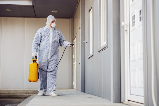 Homme en combinaison de protection contre les virus et masque désinfectant les bâtiments du coronavirus avec le pulvérisateur. Prévention des infections et contrôle de l'épidémie. Pandémie mondiale.