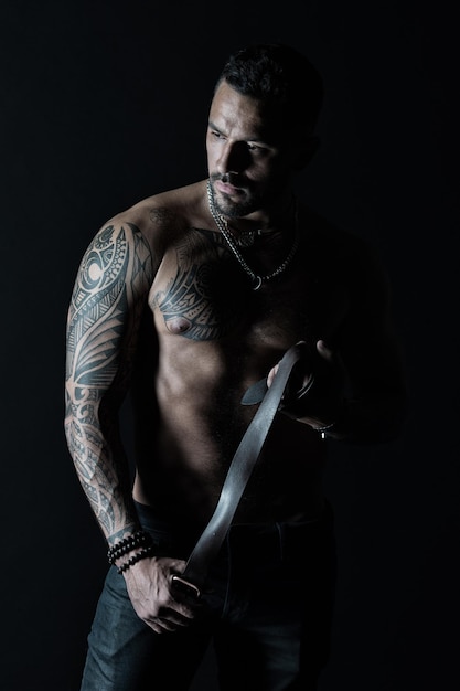 Homme avec conception de tatouage sur la peau. Homme barbu au torse musclé. Mannequin avec ceinture en cuir en jeans. Sportif avec bras et poitrine tatoués. Soins du corps, fitness et sport.