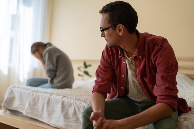 Un homme confus regarde vers une fille qui pleure assise de l'autre côté du lit après une conversation désagréable