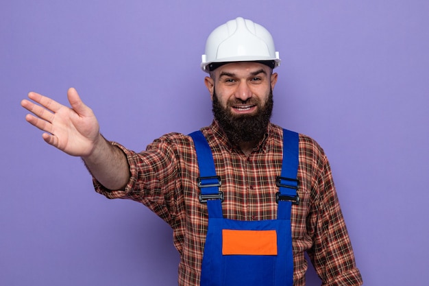 Homme constructeur barbu en uniforme de construction et casque de sécurité regardant la caméra souriant joyeusement levant le bras en agitant la main debout sur fond violet
