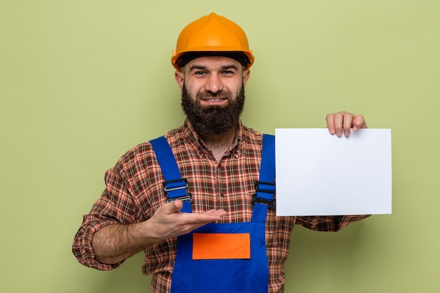 Homme constructeur barbu en uniforme de construction et casque de sécurité tenant une page blanche présentant avec le bras regardant la caméra souriant joyeusement debout sur fond vert