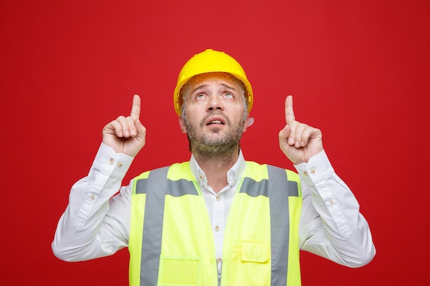 Homme constructeur en uniforme de construction et casque de sécurité levant les yeux confus pointant avec l'index debout sur fond rouge