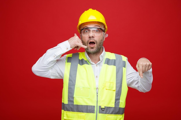 Homme constructeur en uniforme de construction et casque de sécurité portant des lunettes de sécurité regardant la caméra étonné et surpris de faire appelez-moi geste pointant avec l'index vers le bas debout sur fond rouge