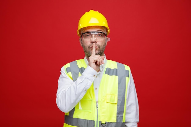 Homme constructeur en uniforme de construction et casque de sécurité portant des lunettes de sécurité regardant la caméra faisant un geste de silence avec le doigt sur les lèvres debout sur fond rouge