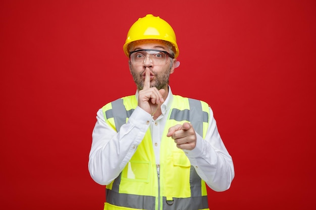 Homme constructeur en uniforme de construction et casque de sécurité portant des lunettes de sécurité regardant la caméra faisant un geste de silence avec un doigt sur les lèvres pointant avec l'index vers la caméra debout sur fond rouge